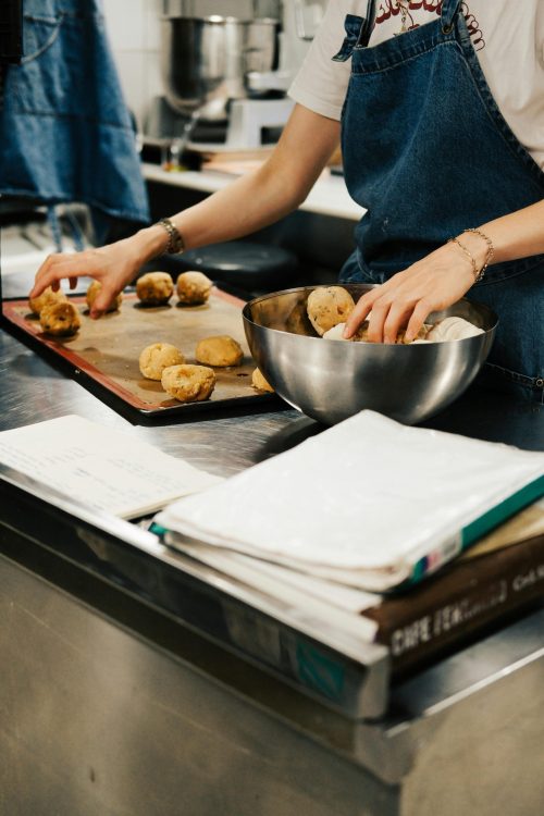 Female chef in denim apron preparing dough balls on a tray in a professional kitchen.